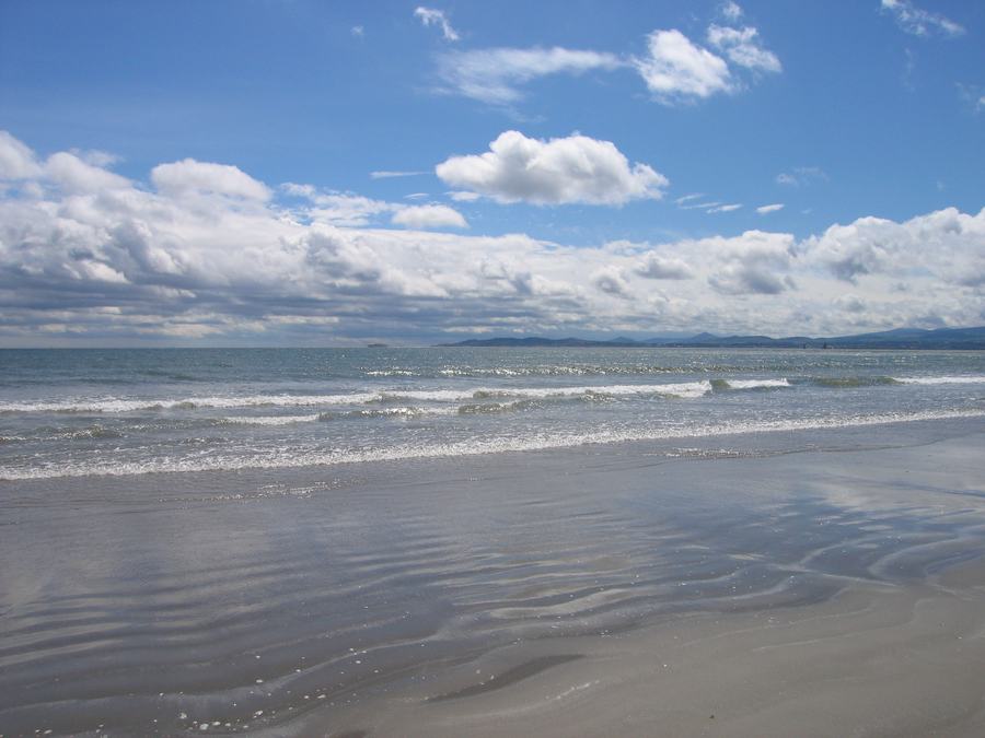 A wide empty sand beach at Dollymount Strand on North Bull Island, Dublin, with breaking waves and hills across the bay.