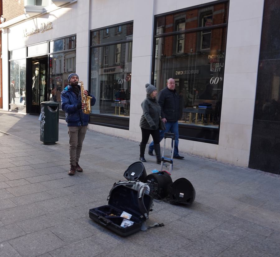 A busker playing a saxophone beside open instrument cases on the pavement of Grafton Street, Dublin, with shoppers walking past a shop window behind him.