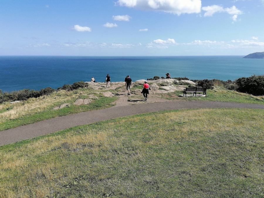 The grassy top of Killiney Hill in County Dublin, with a footpath, a bench and walkers, looking southeast over Killiney Bay and the Irish Sea.