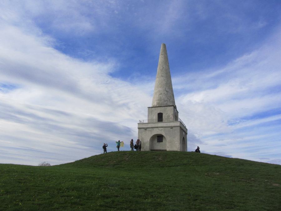 The Mount Mapas obelisk standing on the summit mound of Killiney Hill, County Dublin, with people at its base under a blue sky.