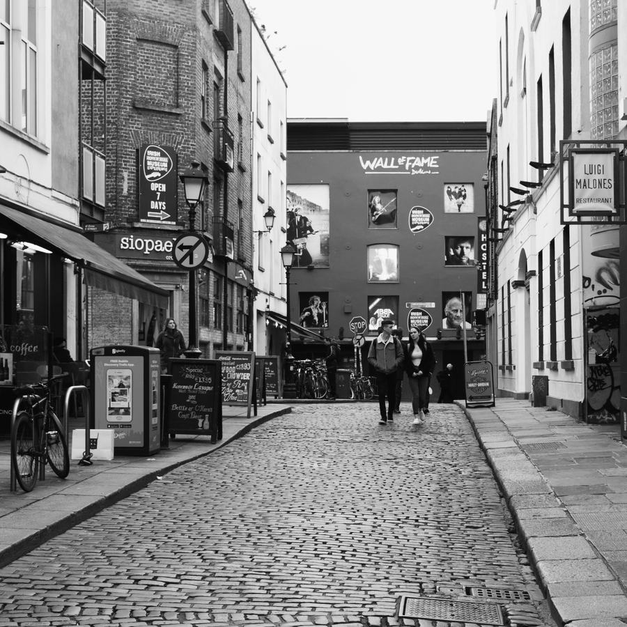 Black and white photograph looking along the cobbles of Cecilia Street in Temple Bar, Dublin, towards a tall wall hung with large portraits of Irish musicians and the logo of the Irish Rock'n'Roll Museum Experience.