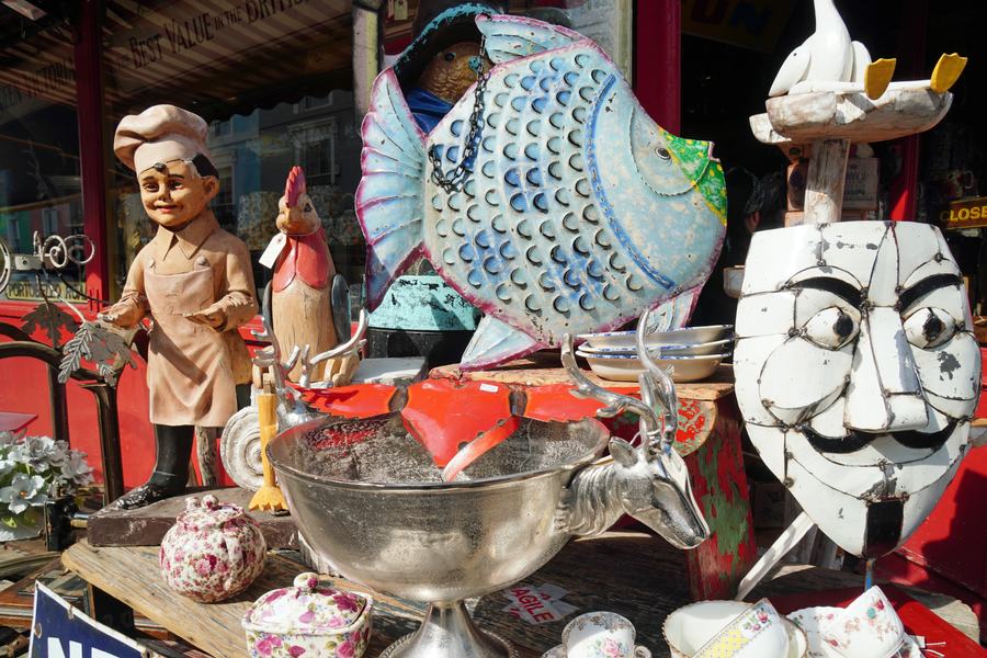 Bric-a-brac stacked outside Alice's Antiques on Portobello Road: a painted fish, a chef figure, a carved mask, silver bowls and china.