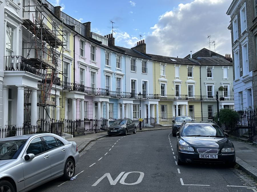 A curving terrace of houses painted pink, cream, blue and yellow on Chalcot Crescent, with cars parked along the kerb.