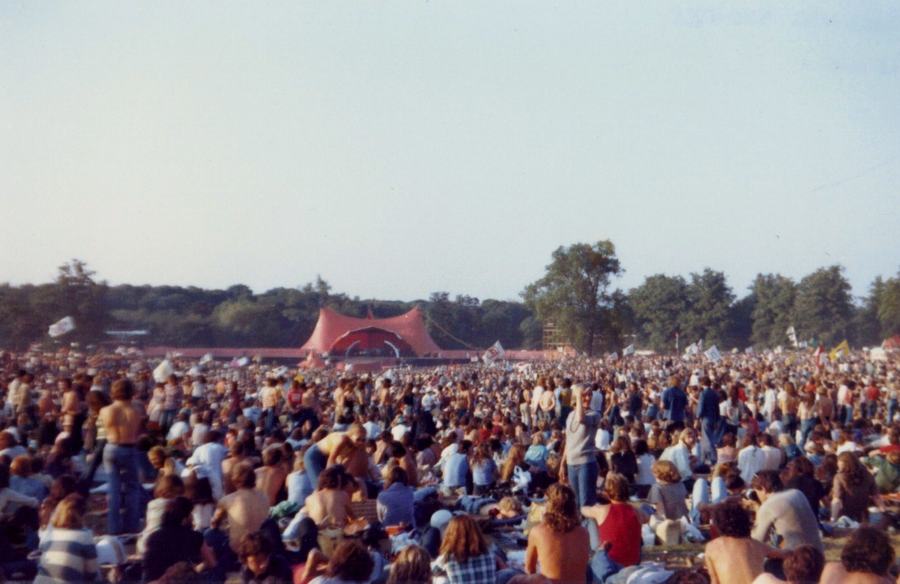A crowd of many thousands sitting on the grass at Knebworth Park in 1976 waiting for the Rolling Stones, a red and white stage in the distance.