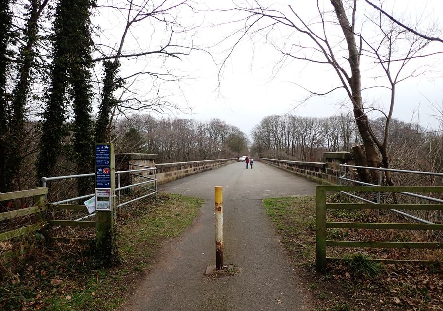 The Nidderdale Greenway cycle path crossing the deck of Nidd Viaduct between bare winter trees, with two walkers in the distance.