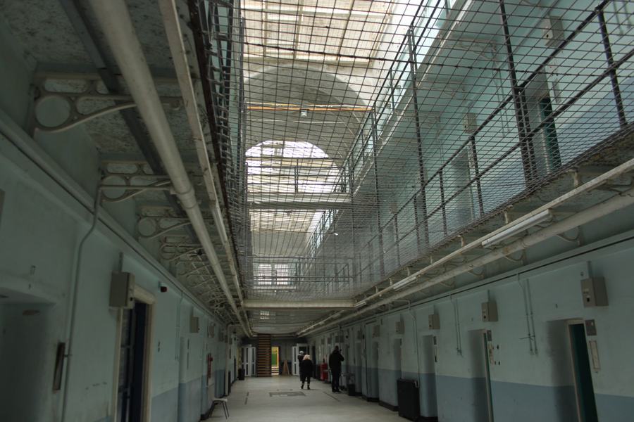 A prison landing inside Shepton Mallet: cell doors along both sides, iron galleries above, anti-suicide netting strung across the well, glazed roof.