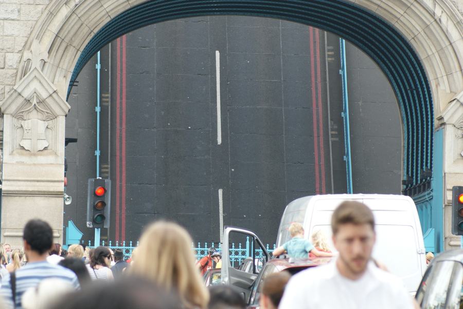 Looking through the arch of a Tower Bridge tower at the raised roadway, with waiting pedestrians and a van in the foreground.