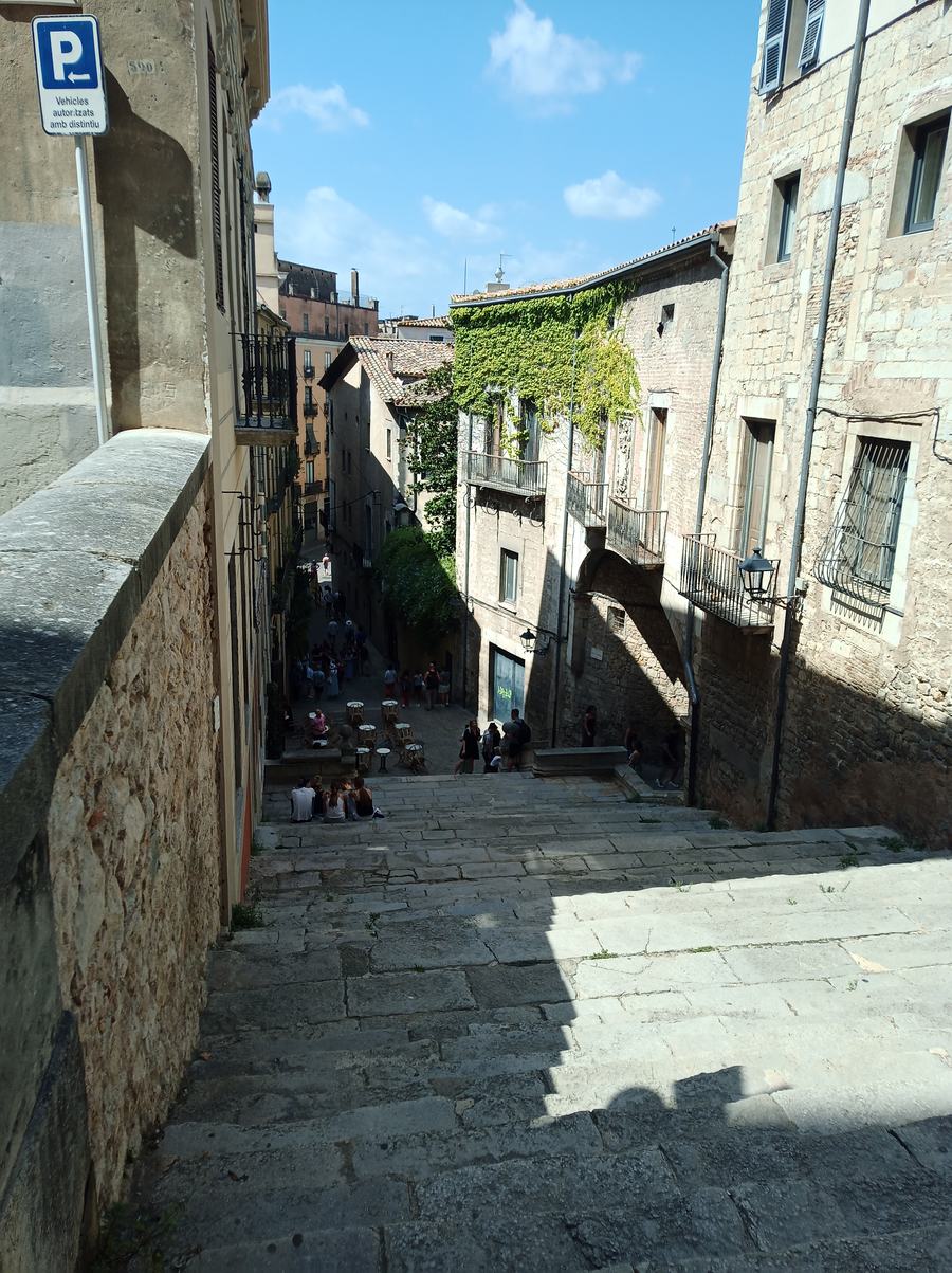 Wide stone steps dropping between tall stone houses, people sitting on the steps, cafe tables at the foot.