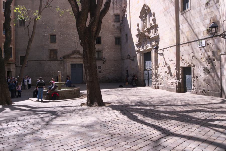 A small enclosed square paved in stone, an octagonal fountain, two large plane trees, a baroque church door on the right, its lower wall pitted.