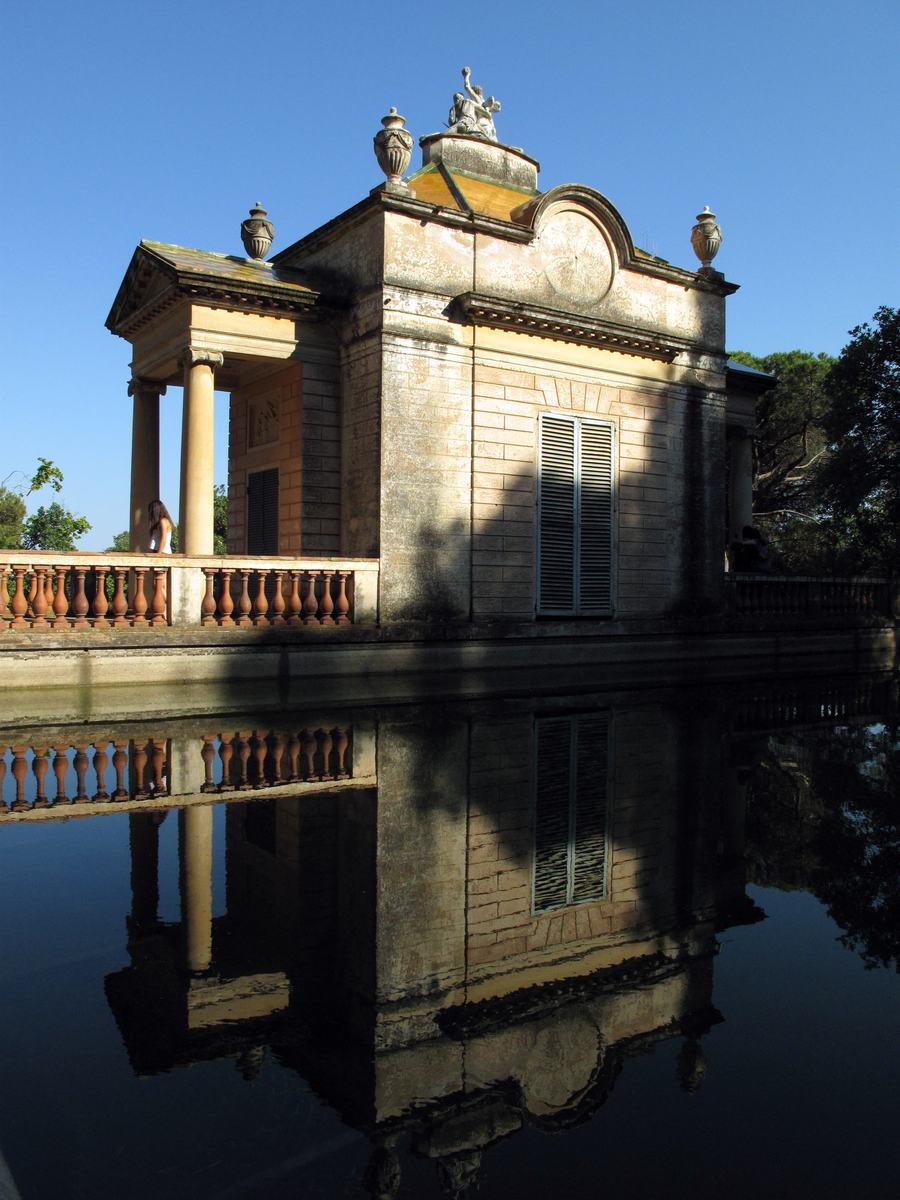 A small columned pavilion with urns on the parapet, mirrored in dark still water.