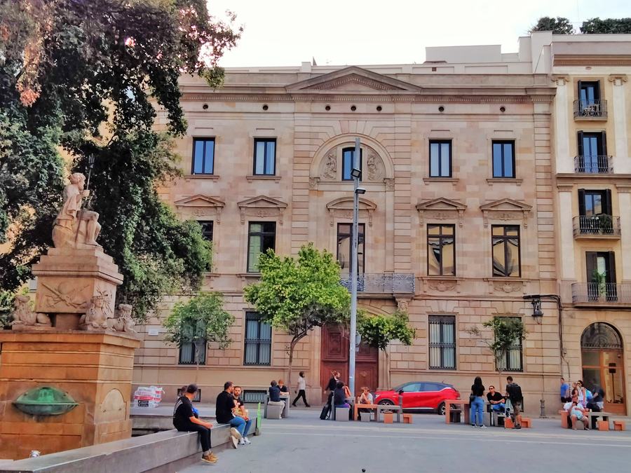A stone fountain with a seated figure at the left, benches with people sitting, a nineteenth-century ashlar facade across the square.