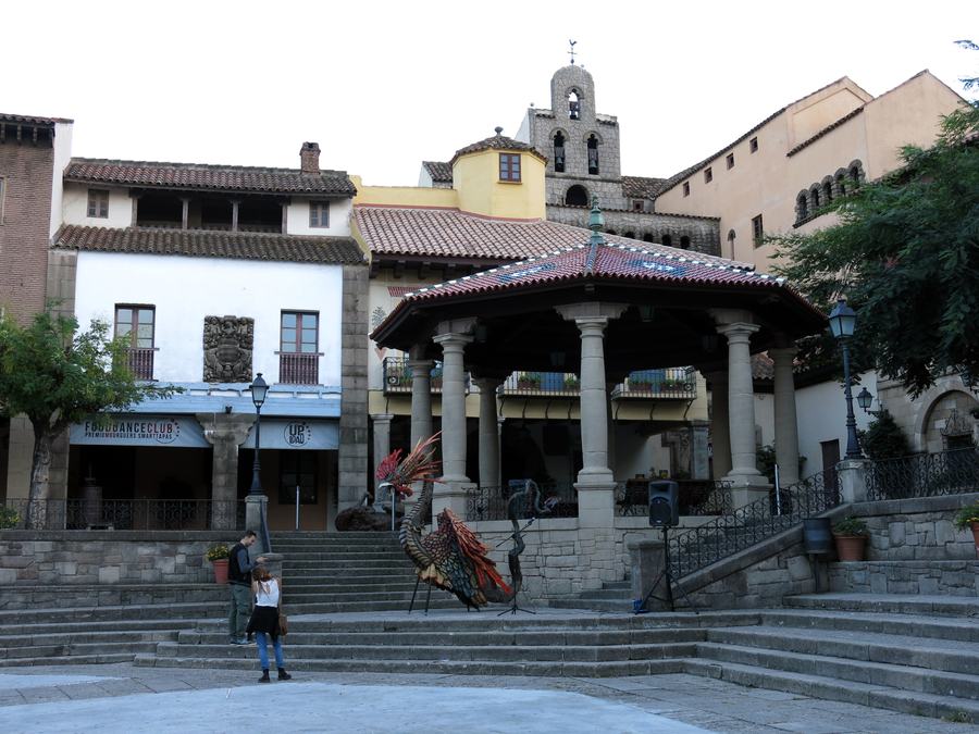 An open square inside the open-air museum, a covered stone pavilion in the middle, a bell tower behind, steps rising on both sides.