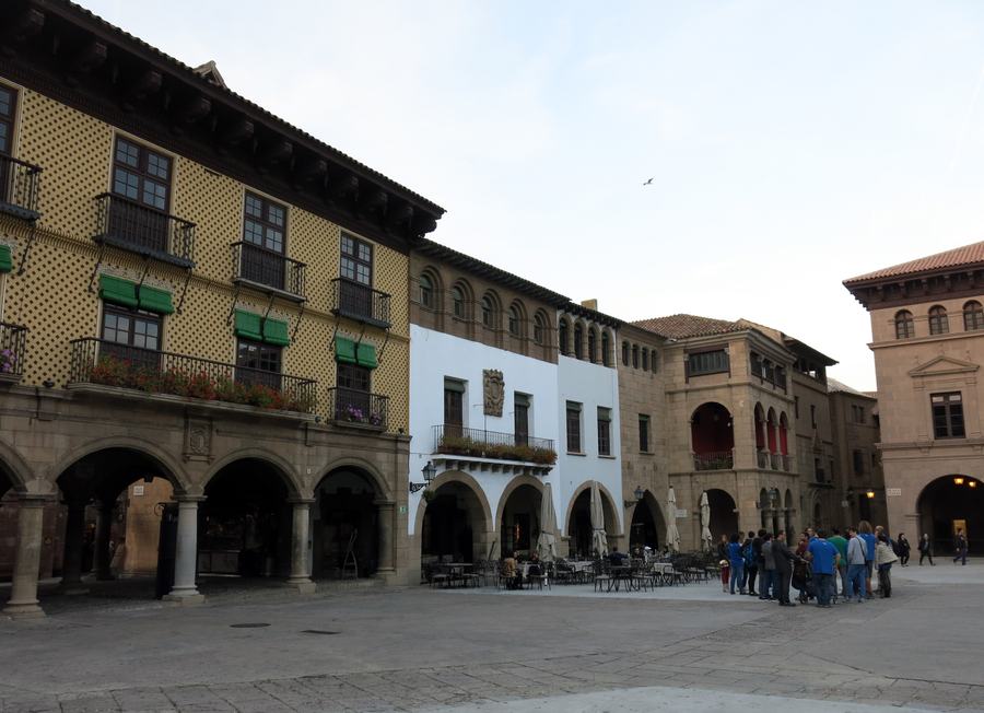 Arcaded buildings around a paved square, a painted timber-balcony house on the left, a group of visitors on the right.