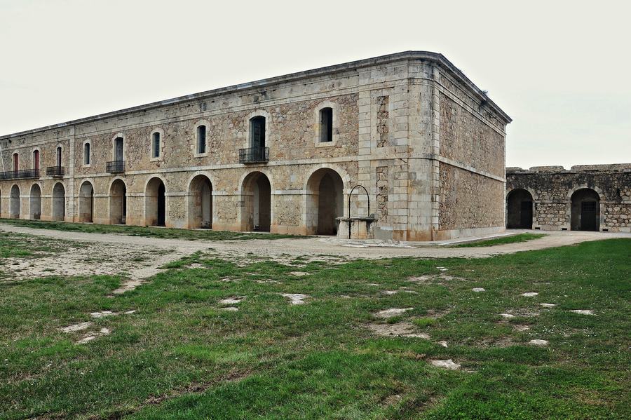 A two-storey arcaded barrack block of pale stone around a grassed courtyard.