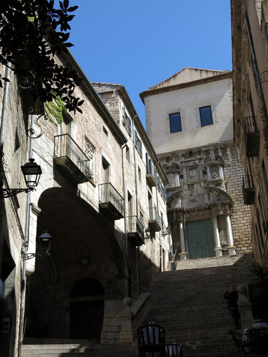 A stone staircase climbing to a baroque church portal, an arched passage under a palace on the left, cafe chairs at the bottom.