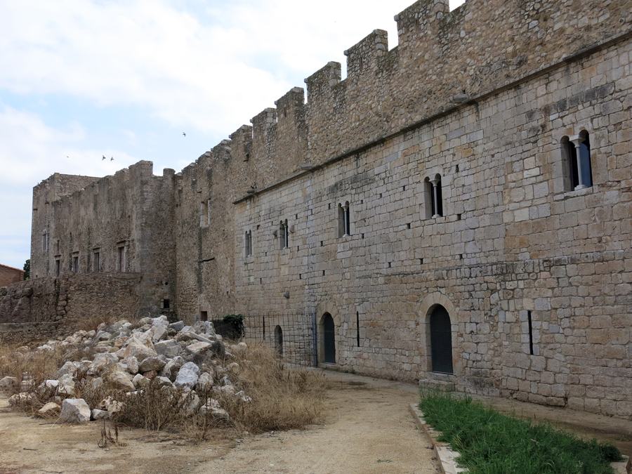 A long crenellated stone range with Romanesque windows and doorways, rubble and dry grass in front.