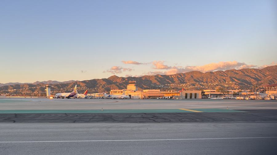 Low airport terminal buildings and a control tower beyond an empty apron, mountains behind