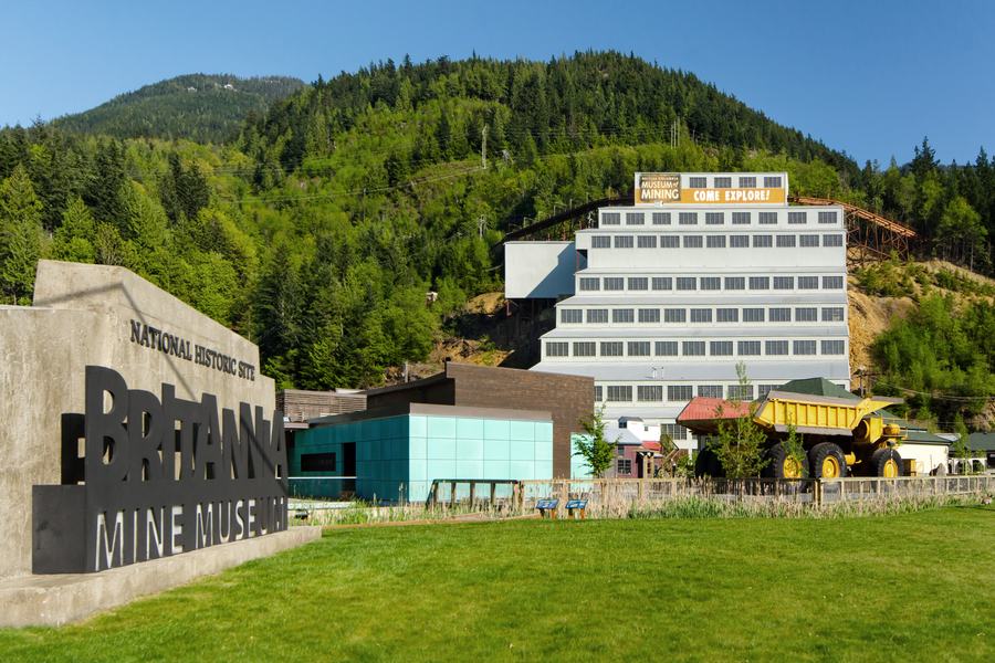 Tall white stepped mill building on a forested hillside above a museum entrance sign that says Britannia Mine Museum, a giant yellow haul truck at right