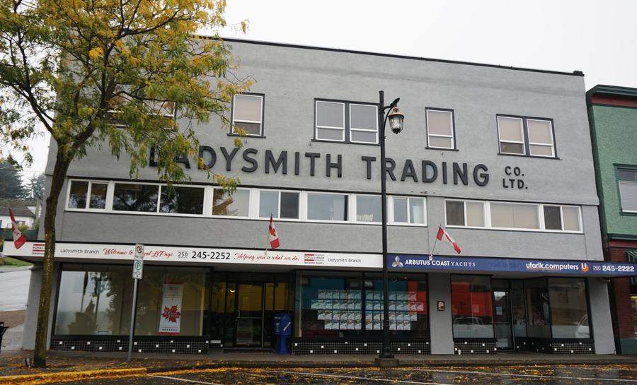 Two-storey grey commercial building with large raised letters reading LADYSMITH TRADING CO. LTD. above shopfronts on a wet main street, a yellowing tree at left