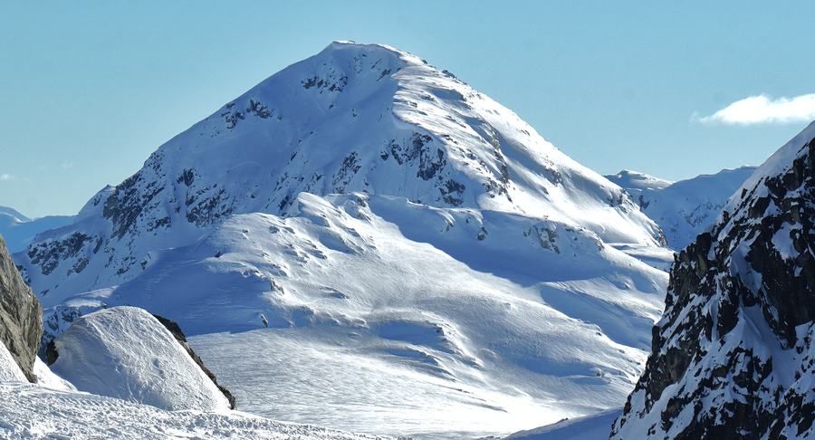 Snow-covered pyramidal peak above a wide snowfield under a clear blue sky, rock wall at right