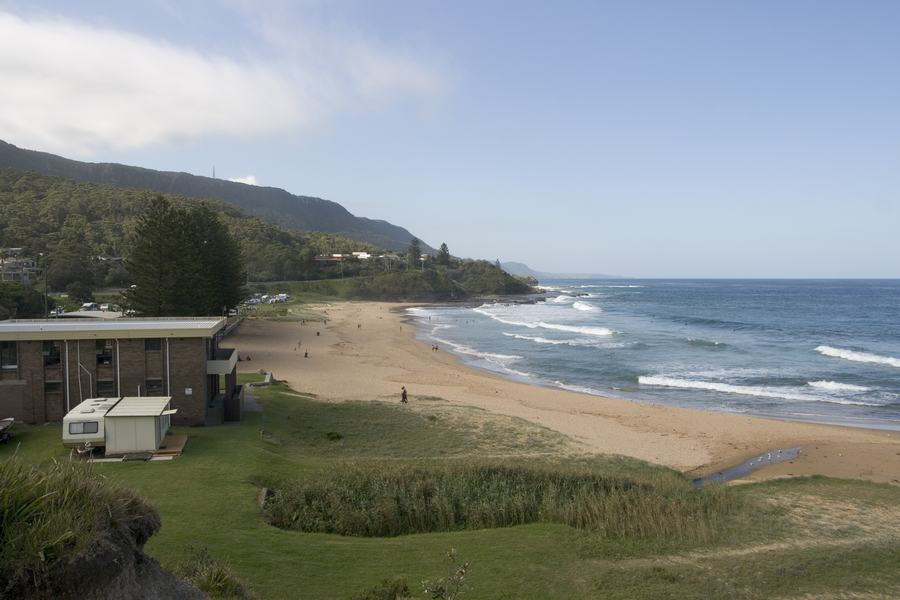 Coledale Beach curving north under the Illawarra escarpment, with a brick building and open grass on the left