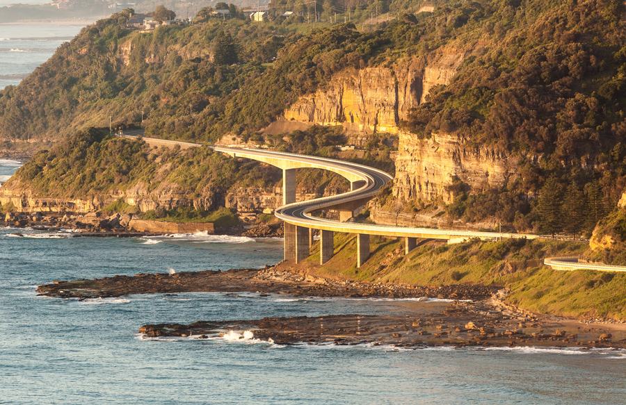 The curving Sea Cliff Bridge runs along the foot of sandstone cliffs above the sea, lit by early morning sun