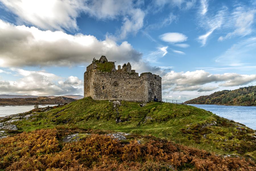 Ruined stone castle on a grassy tidal island with water on either side and bracken in front