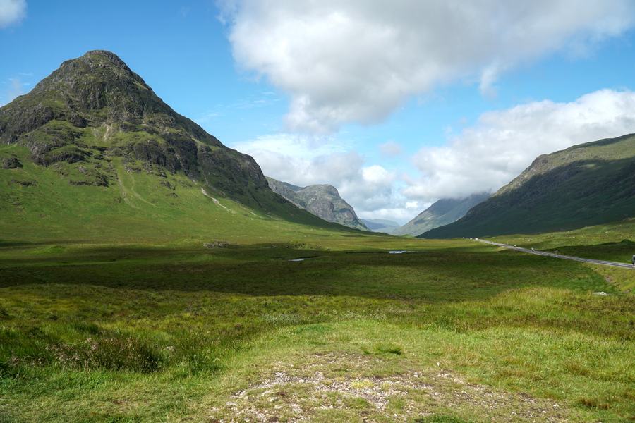 Wide green glen in Glen Coe with a steep conical peak on the left and the A82 road on the right