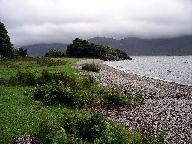 A pebble beach and a wooded promontory on the north shore of Loch Morar under grey cloud