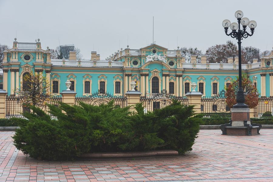 Sunlit turquoise palace facade with white columns, a tall ornate lamp standard and a clipped hedge in front