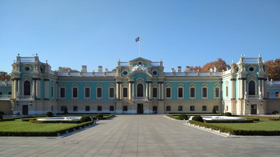 The turquoise and white Mariinskyi Palace behind a wide empty paved forecourt, a Ukrainian flag on the roof