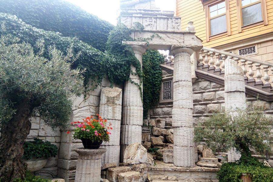 Stone columns and a broken pediment overgrown with ivy, with a potted geranium on the steps