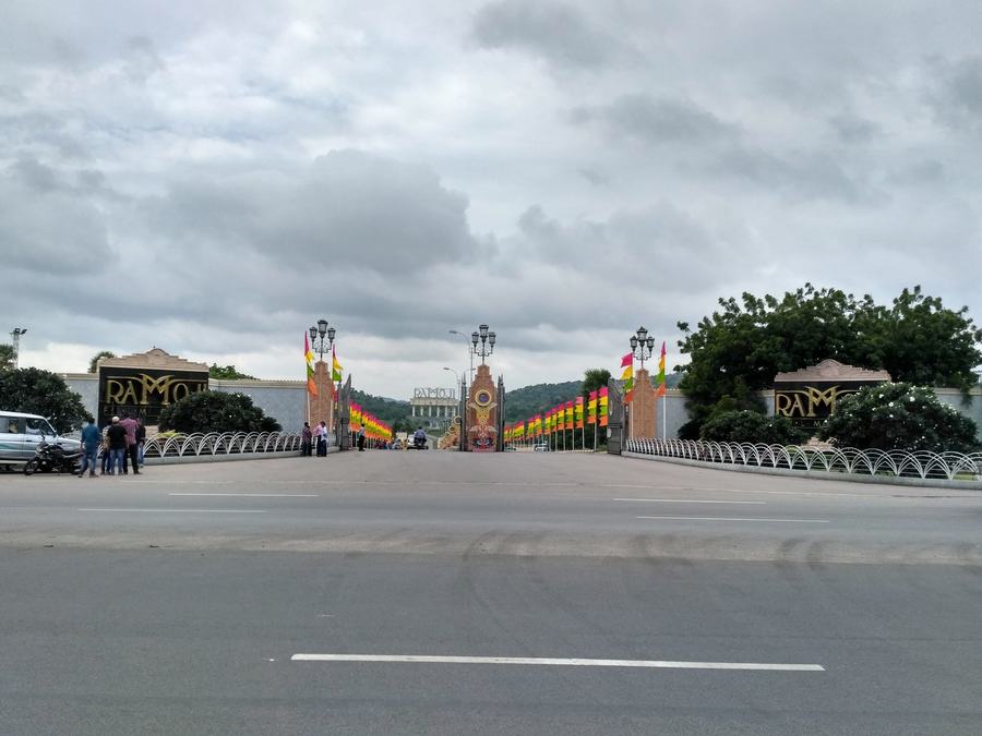 A wide empty road leading to a decorated gateway with flags, hills behind under a grey sky