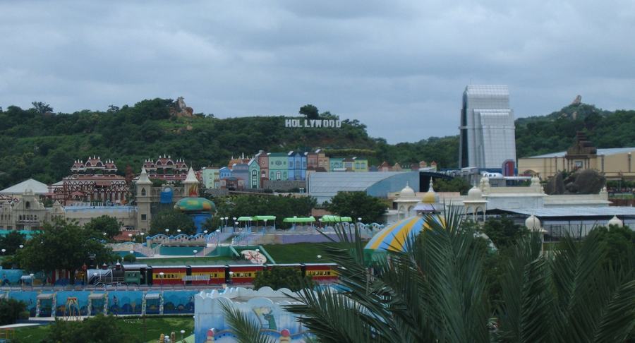 View over a colourful theme park with a small train, and a HOLLYWOOD sign on the green hill behind