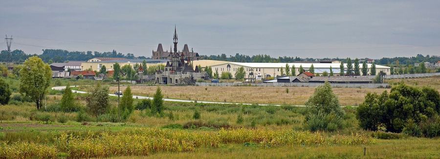 A long low row of pale hangars behind a fence with a small castle-shaped gatehouse in front, seen across rough grassland