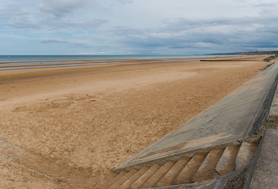 Wide empty sand at Omaha Beach in Normandy with a concrete sea wall and steps in the foreground