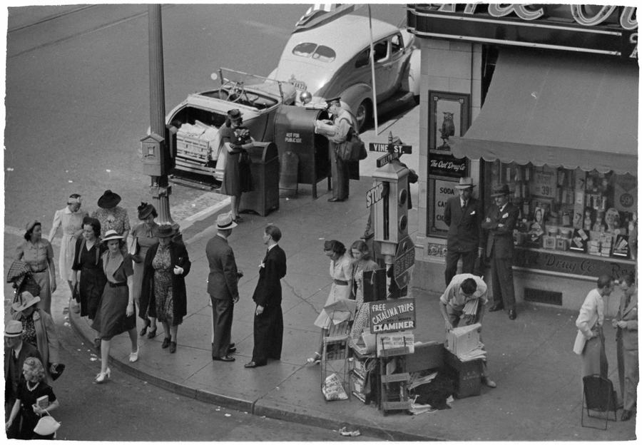 Black and white overhead view of pedestrians in 1940s clothes on a street corner beside a drug store window
