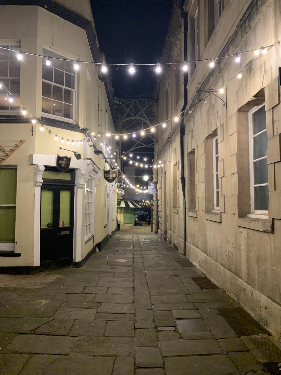 Narrow paved lane at night strung with fairy lights between old buildings, Bristol