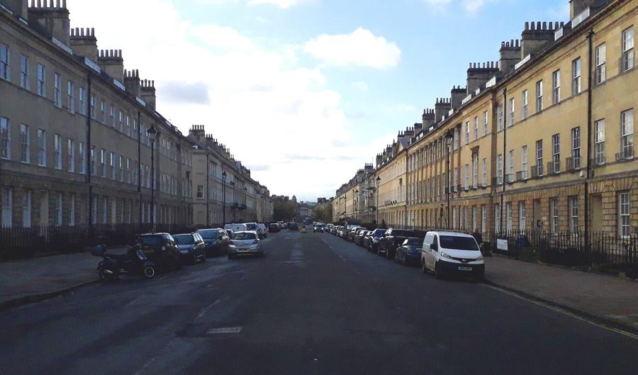 Very wide straight Georgian street lined with uniform stone terraces and parked cars, Bath