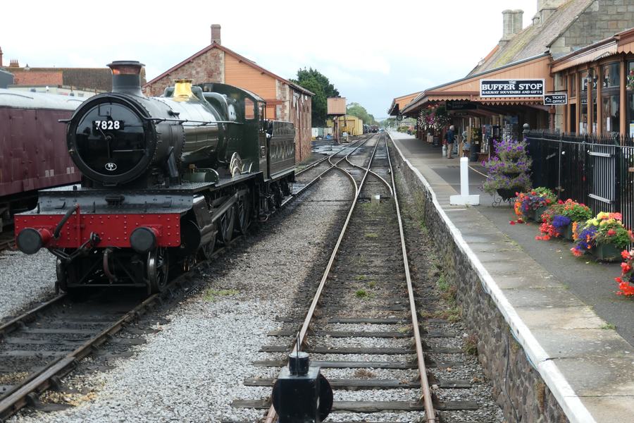 Steam locomotive beside a long platform with a canopy and flower baskets at Minehead station