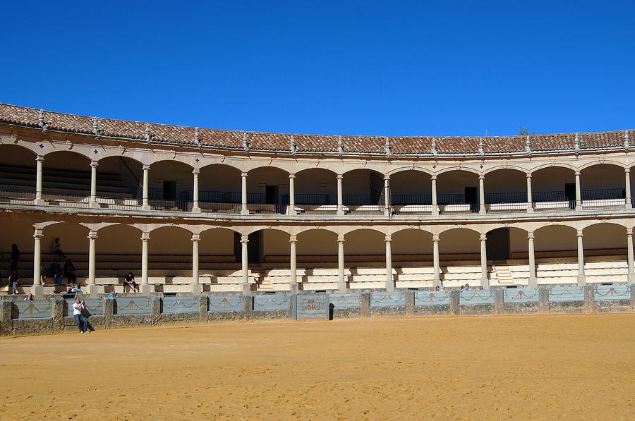 Empty sandy bullring surrounded by two tiers of stone arcades with tiled roof under a blue sky, Ronda