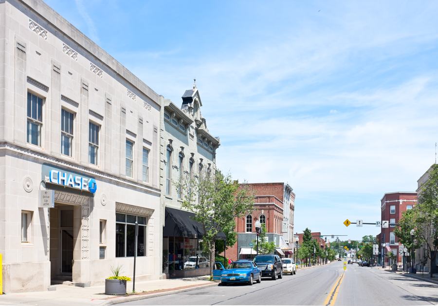 A small-town main street of low brick and stone commercial buildings, cars parked at an angle, seen looking down the road.