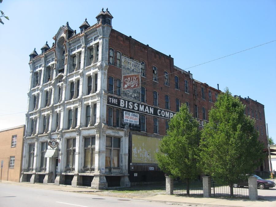 A tall five-storey red brick corner building with peeling white paint on the front and a faded painted sign reading THE BISSMAN COMP on the side.