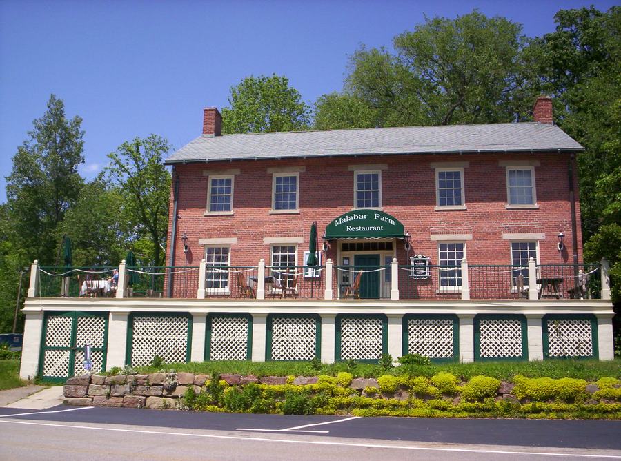 A two-storey white and red clapboard building with a green awning and a low hedge, beside a country road.