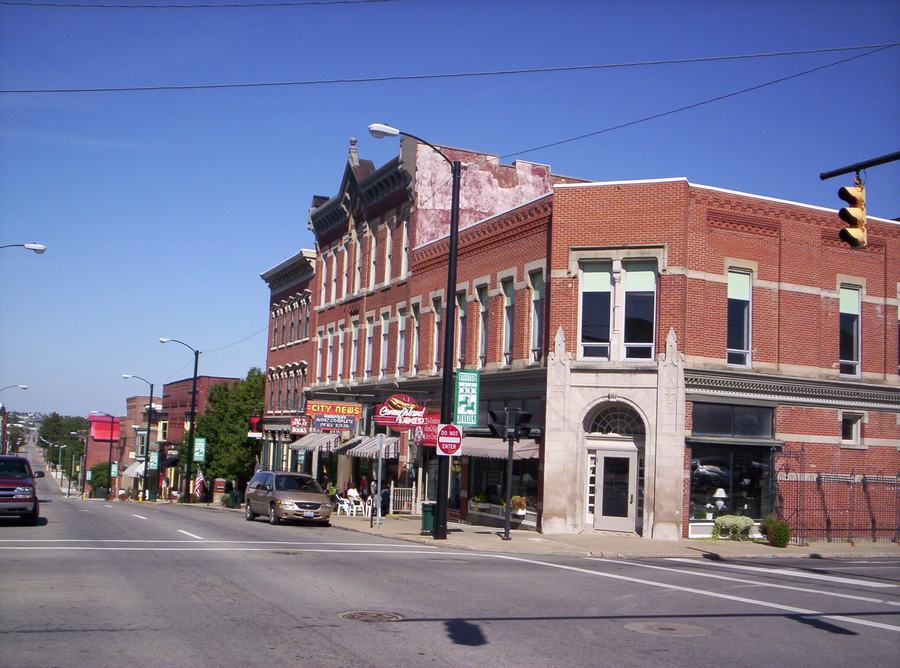 A downtown street of two- and three-storey brick storefronts with awnings and traffic lights, cars parked at the kerb.