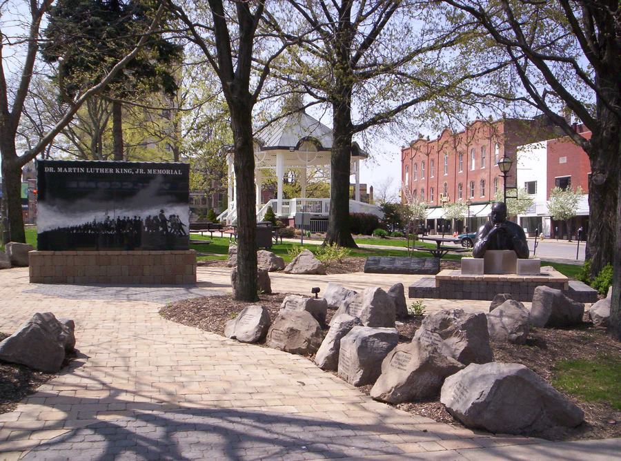 A city park with mature trees, a paved path, boulders and a low memorial wall, brick downtown buildings visible through the trees.