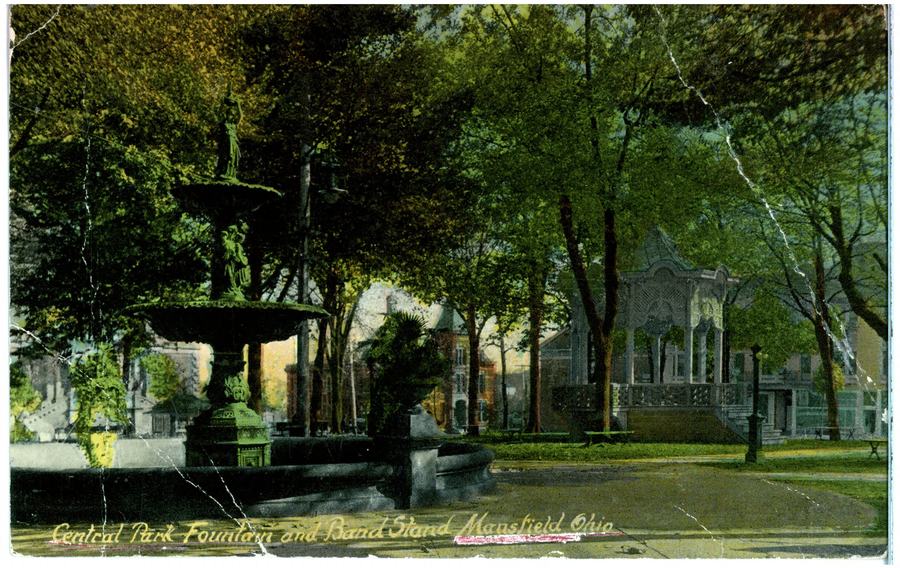 A colourised early twentieth century postcard of a wooded city park with a tiered stone fountain on the left and an open wooden band stand on the right.