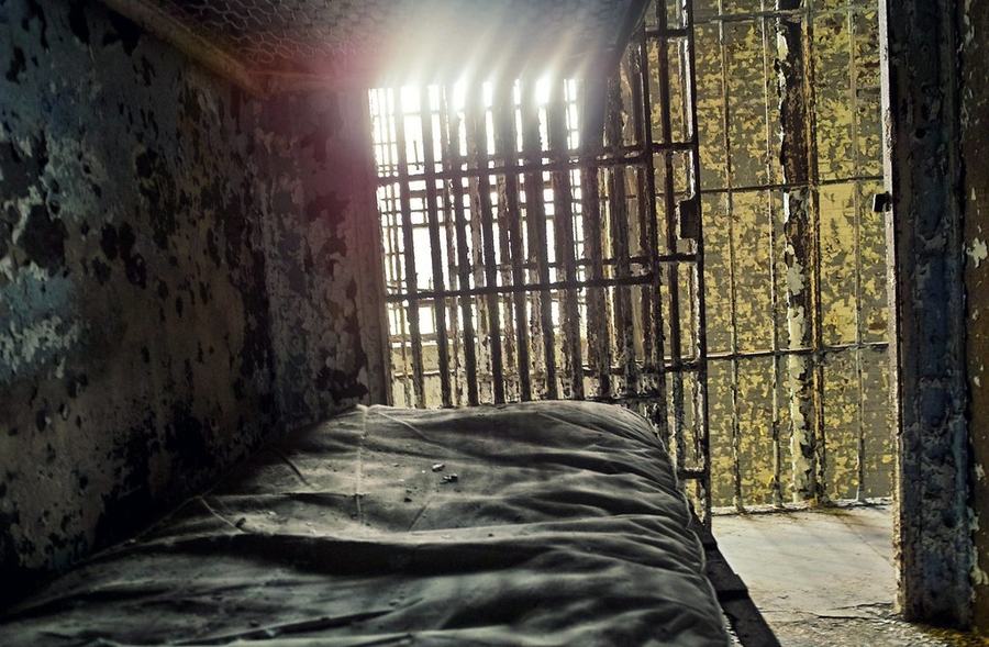 A narrow prison cell with a stripped bed, rusting walls and a barred window letting in bright light.