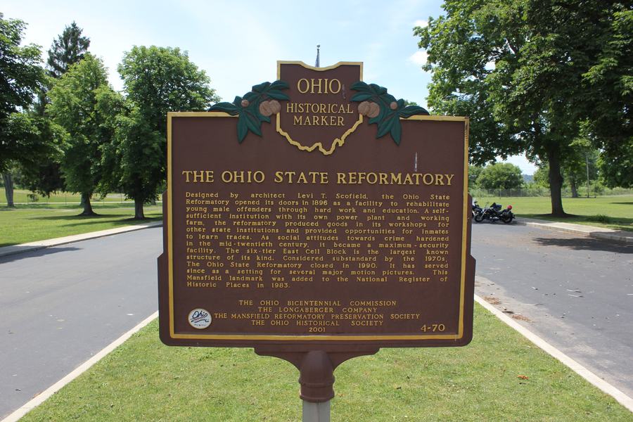 A brown and gold cast metal marker on a post beside a driveway, headed OHIO HISTORICAL MARKER and THE OHIO STATE REFORMATORY.