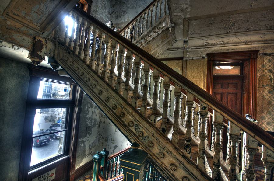An ornate iron and stone staircase with a carved balustrade inside a decaying institutional building, wooden doors at the top.